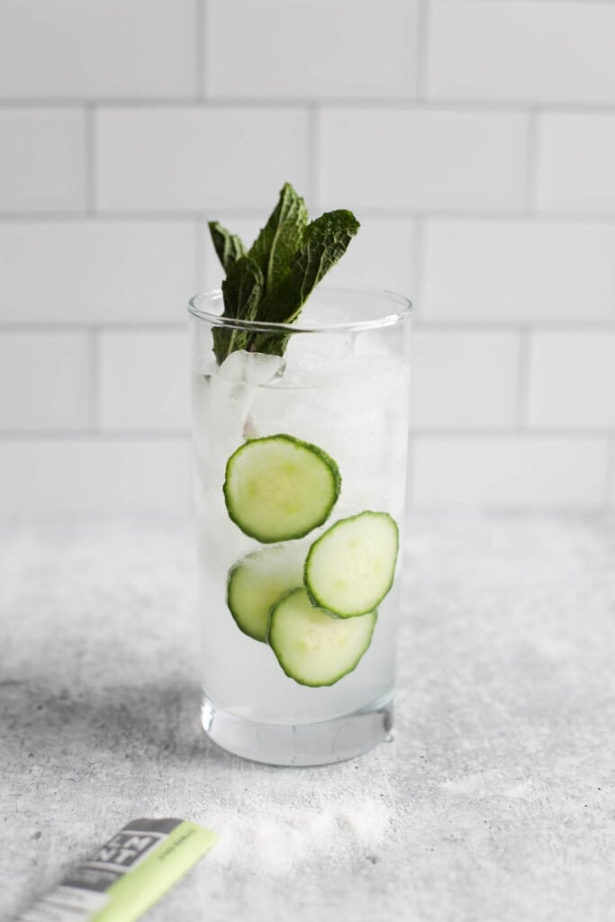 Citrus Salt LMNT Electrolyte Drink Mix in a glass of ice water, garnished with fresh cucumbers and mint. 