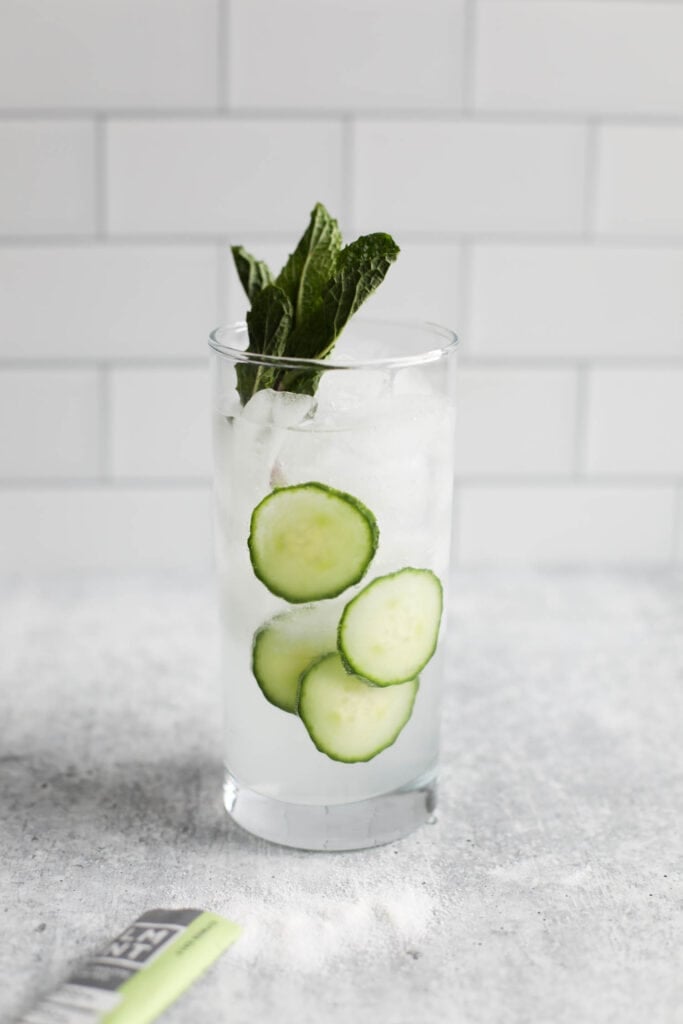 Tall clear glass filled with ice and electrolyte water with floating cucumber slices and a sprig of mint against a white tiled background