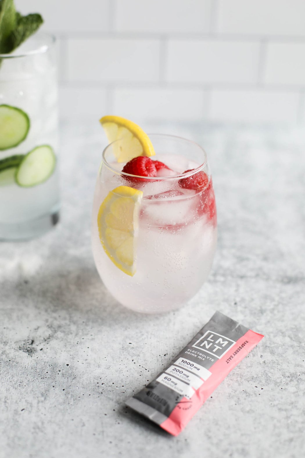 A glass of water and LMNT Raspberry Salt garnished with ripe, fresh raspberries, and lemon slices. 