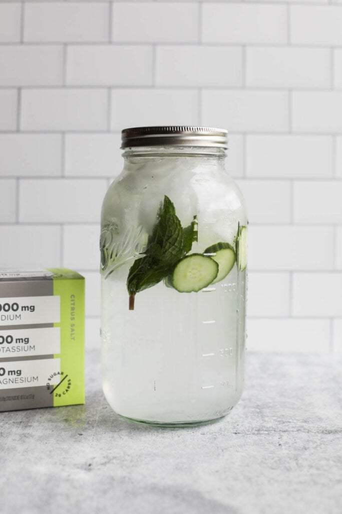 A 64 oz mason jar full of ice water and LMNT Electrolyte Drink mix, garnished with fresh cucumbers and mint. 