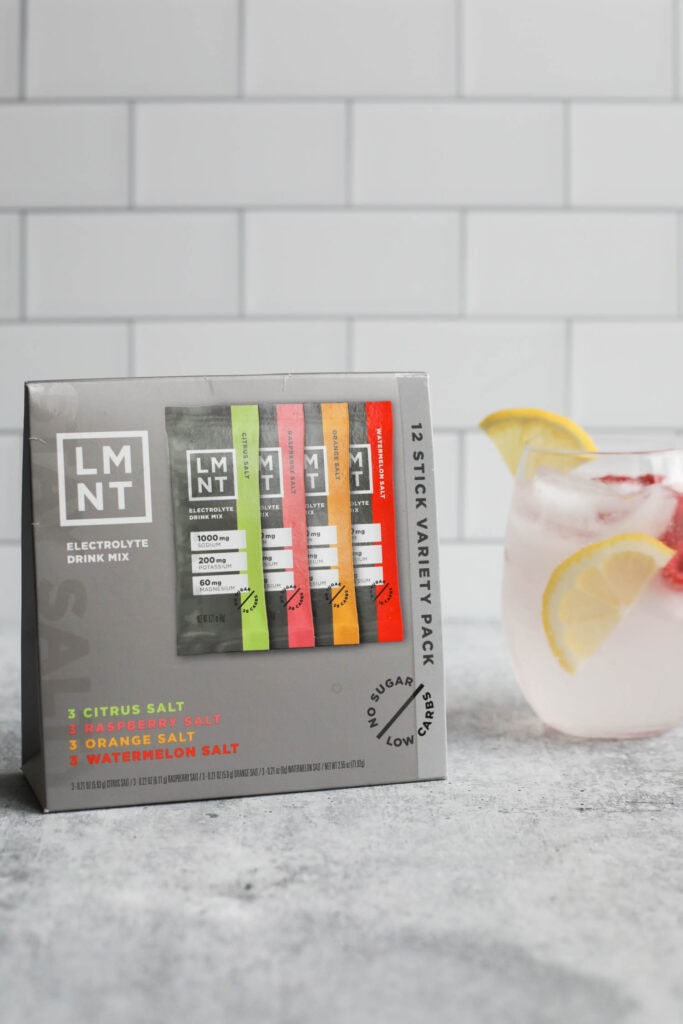 A variety box of LMNT Electrolyte Drink Mix that includes Citrus salt, Raspberry Salt, Orange Salt, and Watermelon Salt, with a glass of LMNT Electrolyte Drink Mix and water next to the box. 