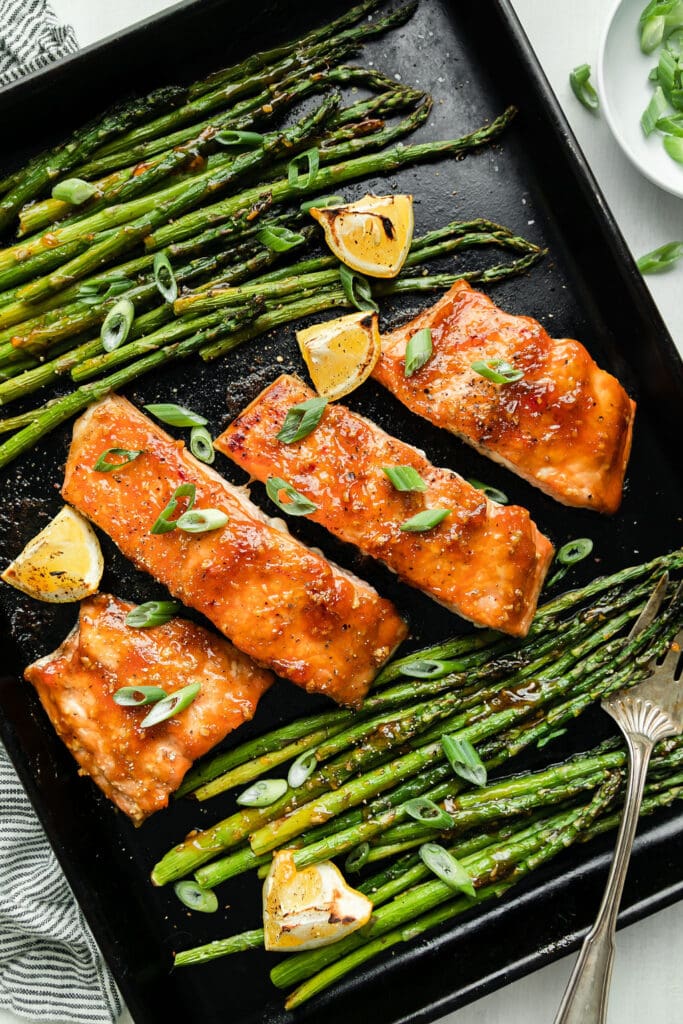 Oven baked salmon filets coated with honey glazed sauce on sheet pan next to roasted asparagus and lemon wedges.