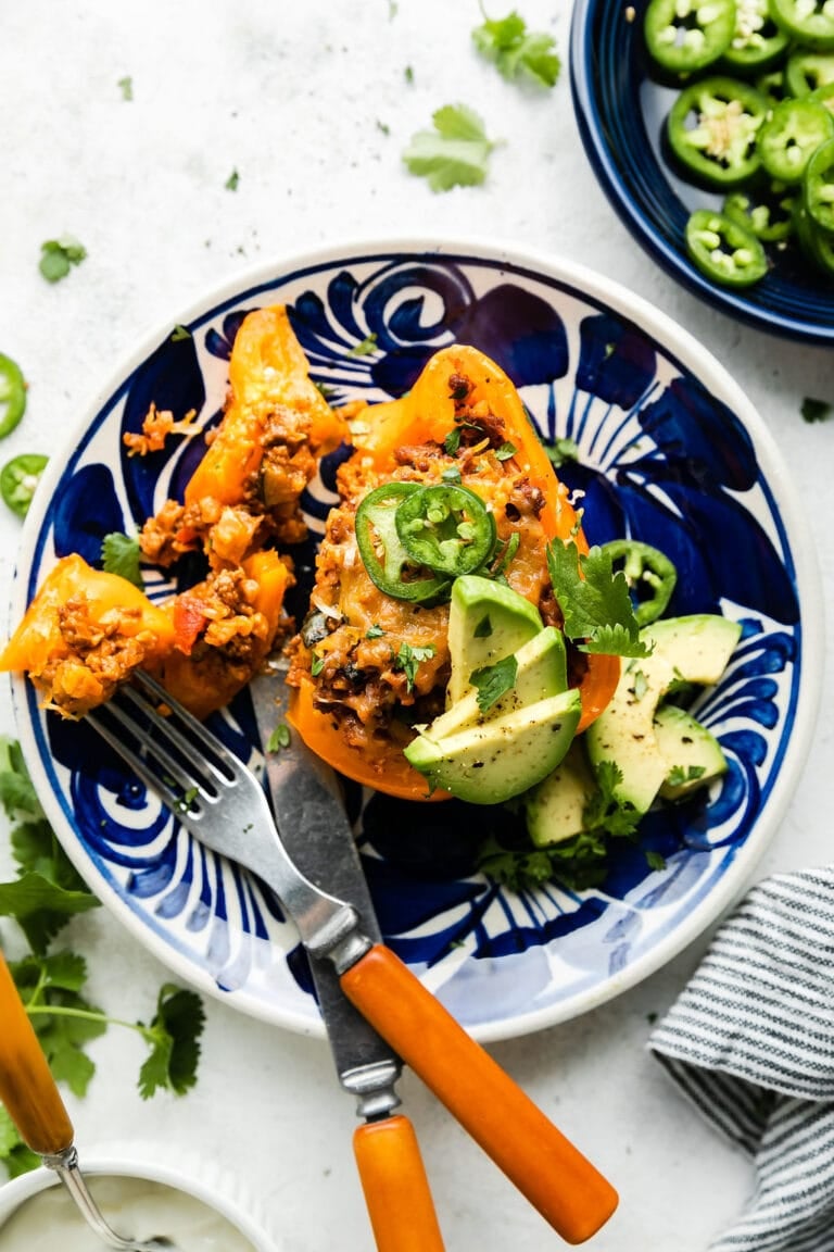 A blue plate with a fork and knife cutting into a yummy stuffed pepper topped with melted cheese, avocado, and jalapeno. 