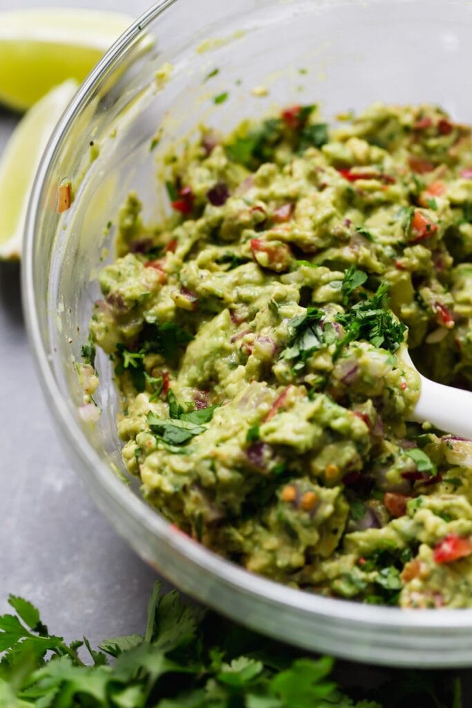 Homemade guacamole being stirred with white spatula in clear mixing bowl