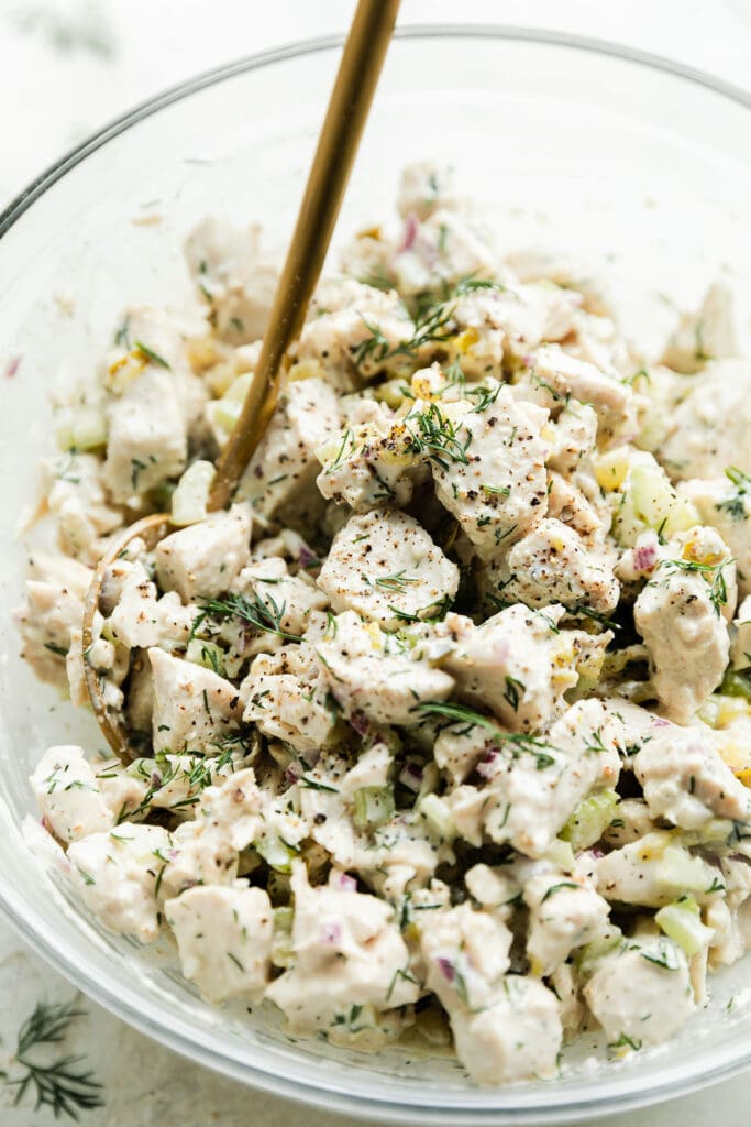 Close up view dill pickle chicken salad in mixing bowl sprinkled with fresh dill and cracked black pepper.