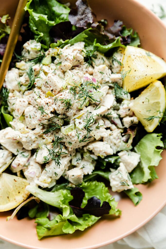 Close up view dill pickle chicken salad served over mixed greens in a pink bowl with lemon wedges on side.