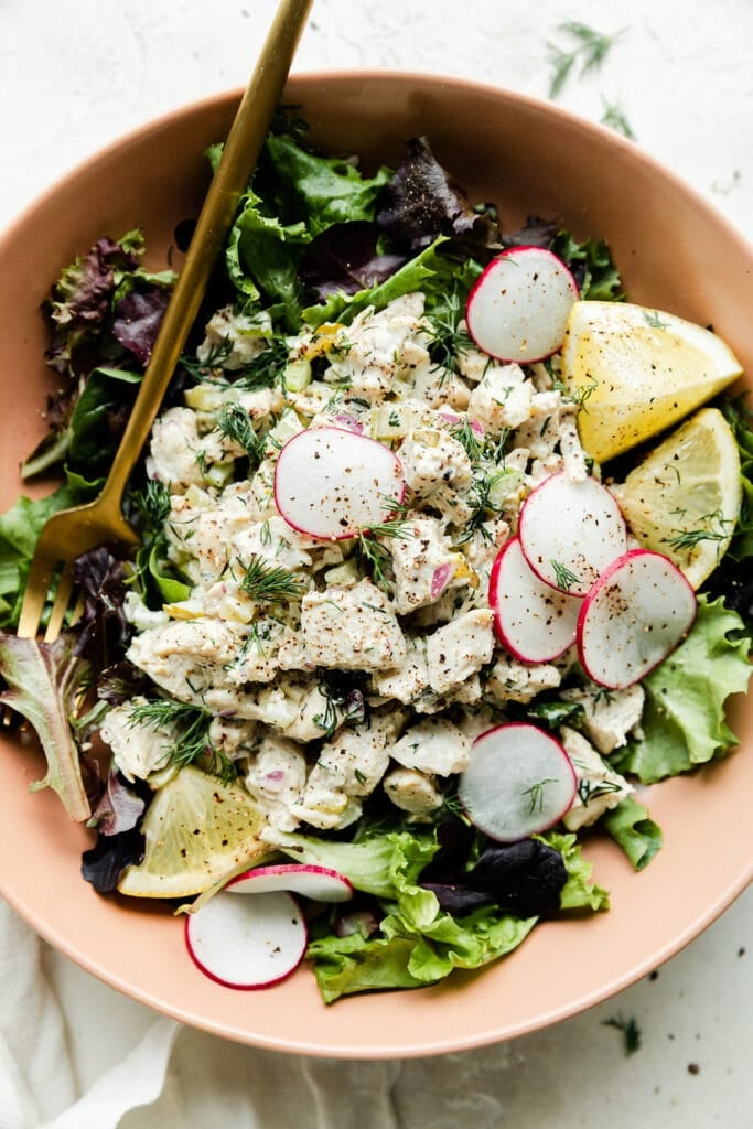 Overhead view dill pickle chicken salad served over mixed greens in a bowl with gold cutlery.