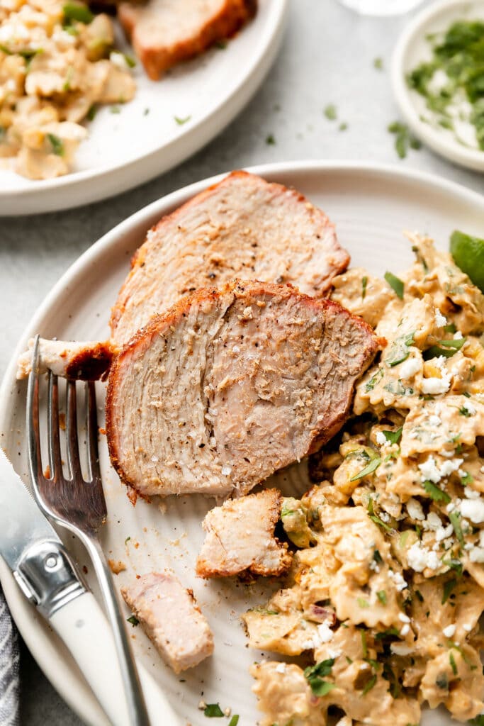 Close up view serving of grilled pork loin with easy rub on stone plate with side of Mexican street corn pasta salad.