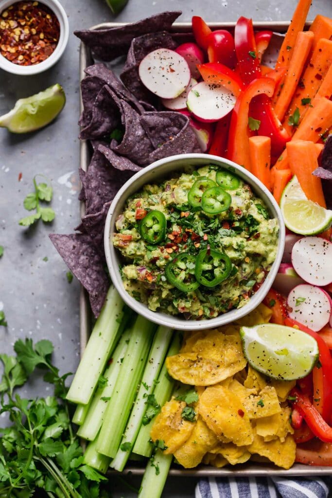 Stone bowl filled with chunky homemade guacamole topped with jalapeno slices, on tray with colorful veggies and blue chips