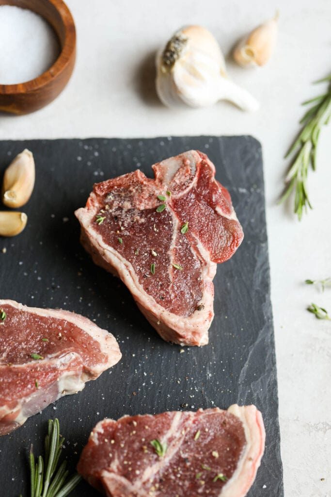 Raw lamb chops seasoned with sea salt and fresh herbs on black cutting board