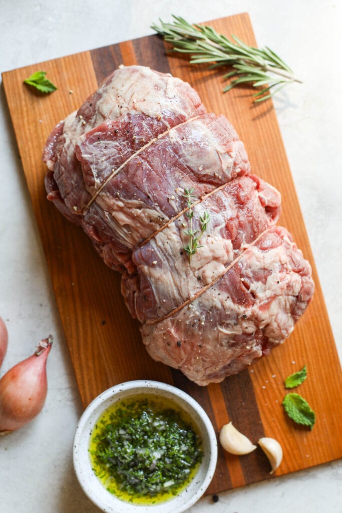 Raw lamb roast wrapped in twine on cutting board with fresh rosemary sprigs.