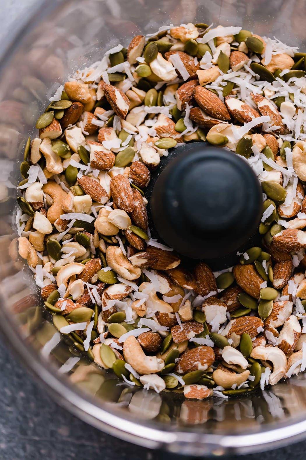 Food processor bowl filled with almonds, cashews, pepitas, and coconut shreds