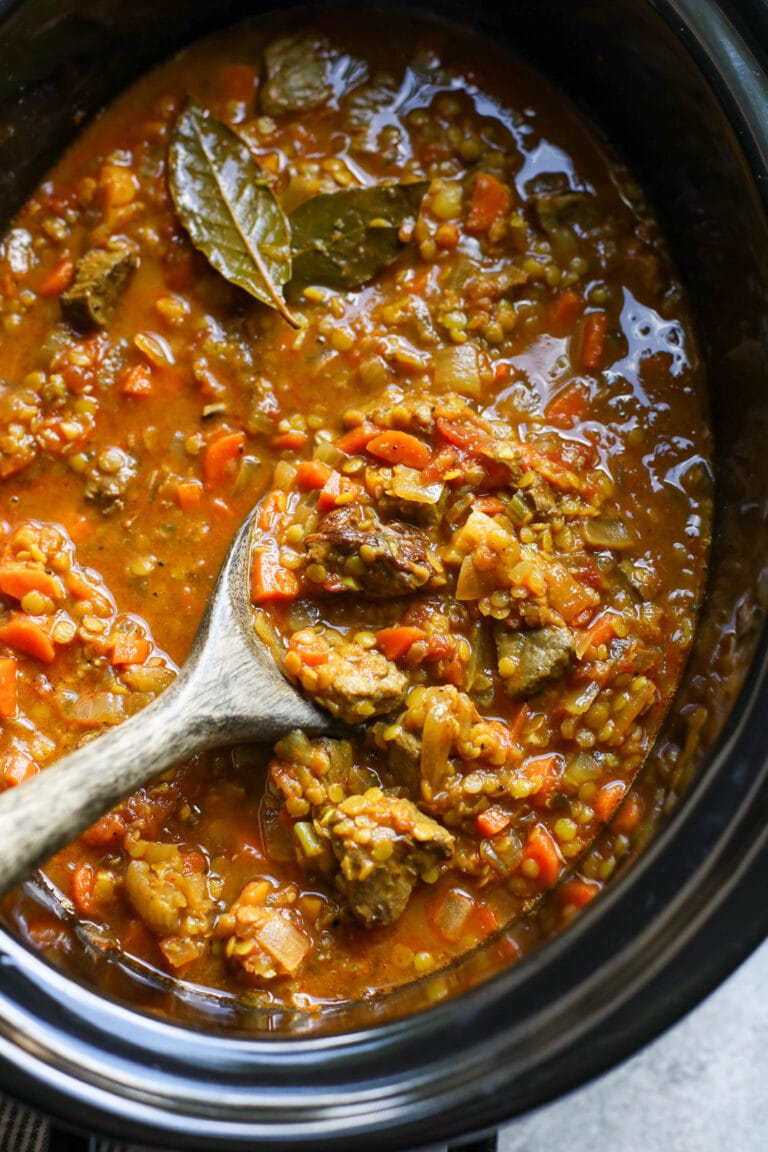 Overhead view of a crockpot full of beef and lentil stew being scooped with a wooden spoon. 