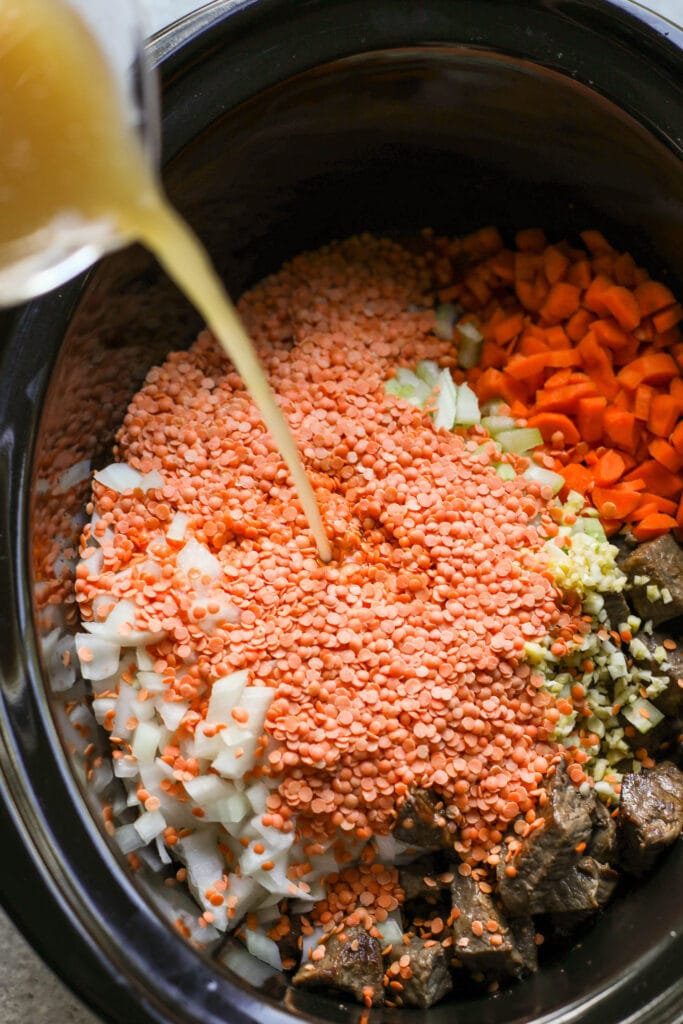Broth being poured over red lentils and ingredients for beef and lentil stew in black slow cooker