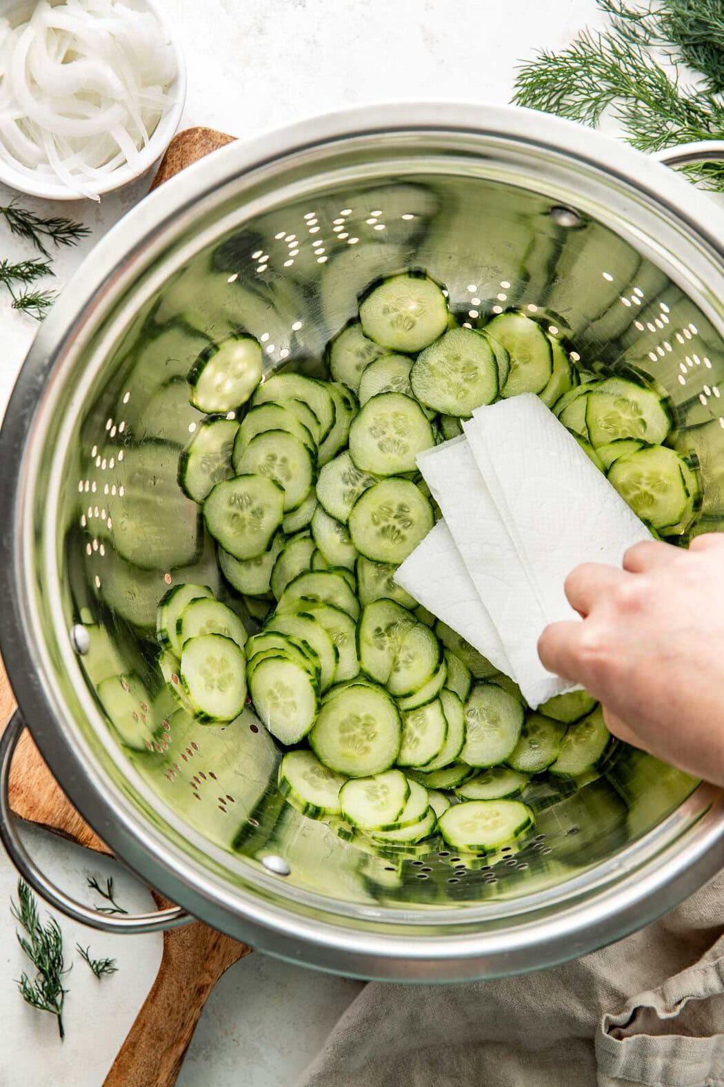 Overhead view of sliced cucumbers in a stainless-steel strainer with a hand using a paper towel to soak up liquid.