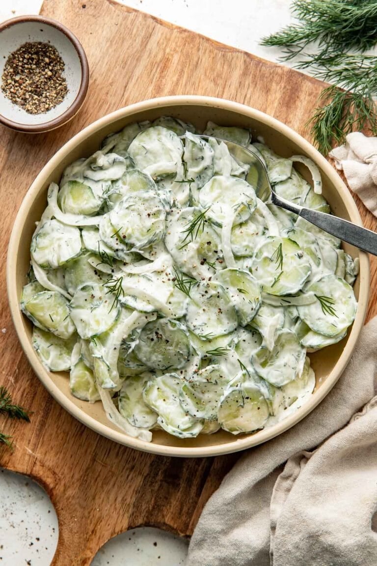 Overhead view of a bowl of creamy cucumber salad sitting on top of a cutting board and with a serving spoon dipped into the salad.