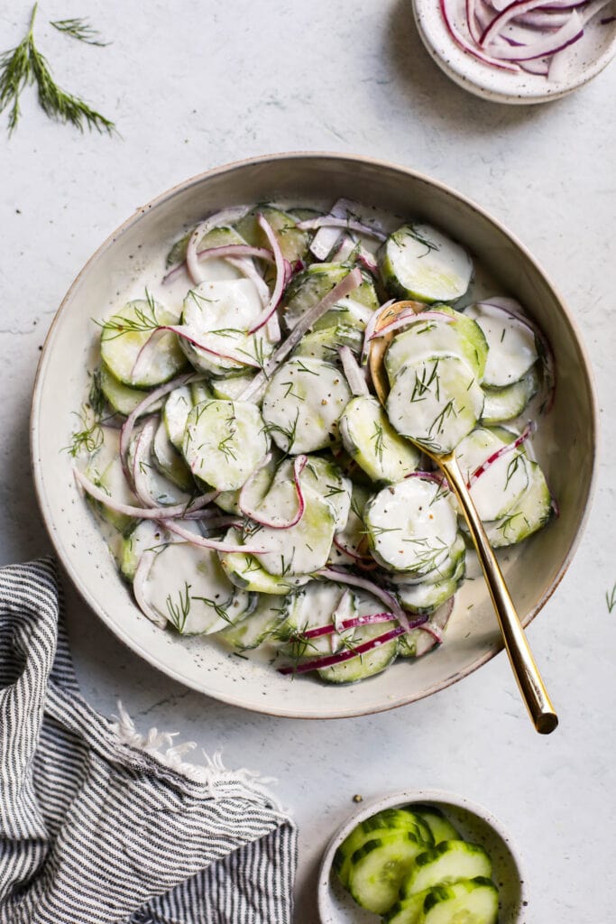 A shallow stone bowl filled with creamy cucumber salad, a gold serving spoon in salad