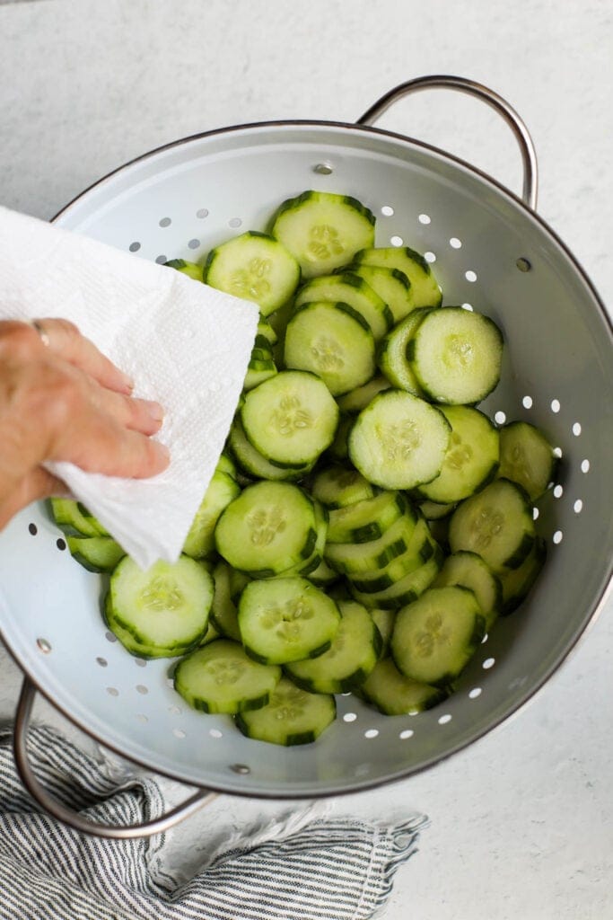 A paper towel patting cucumber slices dry in a white colander