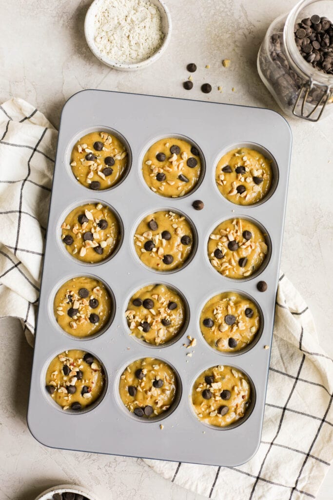 Overhead view protein muffin batter in muffin tin topped with chocolate chips and coconut shreds