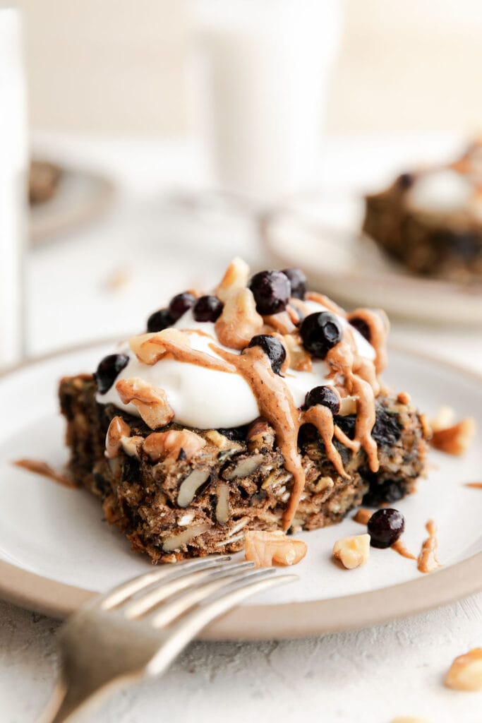 A serving of oatmeal breakfast bars with blueberries on white plate topped with yogurt, almond butter, frozen blueberries, and walnuts
