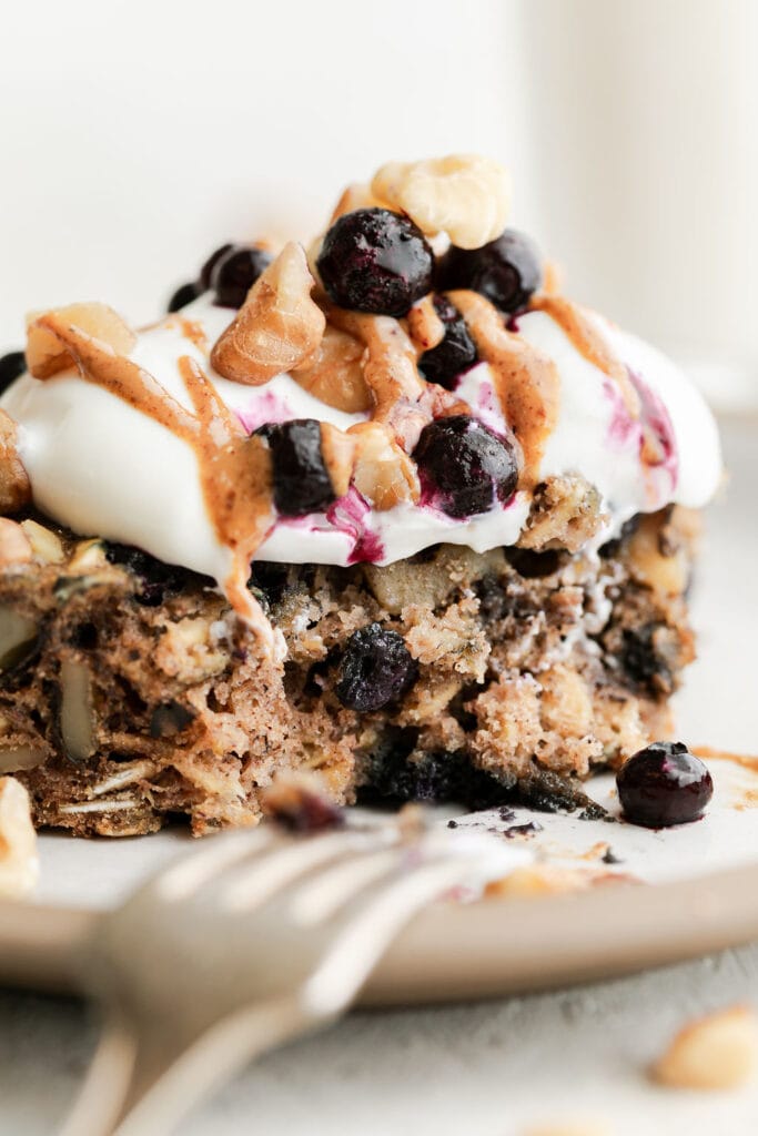 Side view serving of oat breakfast bar with walnuts and wild blueberries on plate topped with yogurt, frozen wild blueberries and walnuts