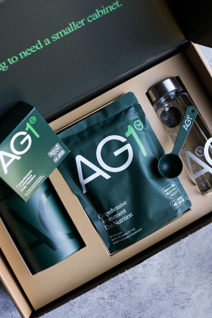 An opened box of AG1 supplements showing the branded water bottle, supplement pouch, and container