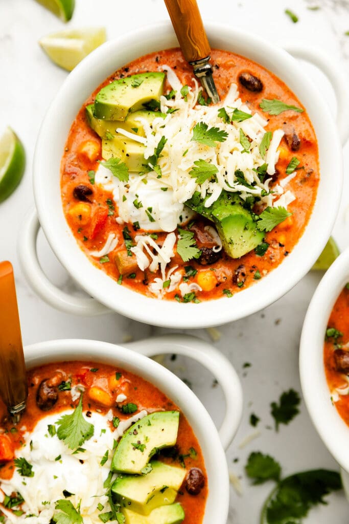 A white bowl of taco soup topped with avocado, cheese, and cilantro.