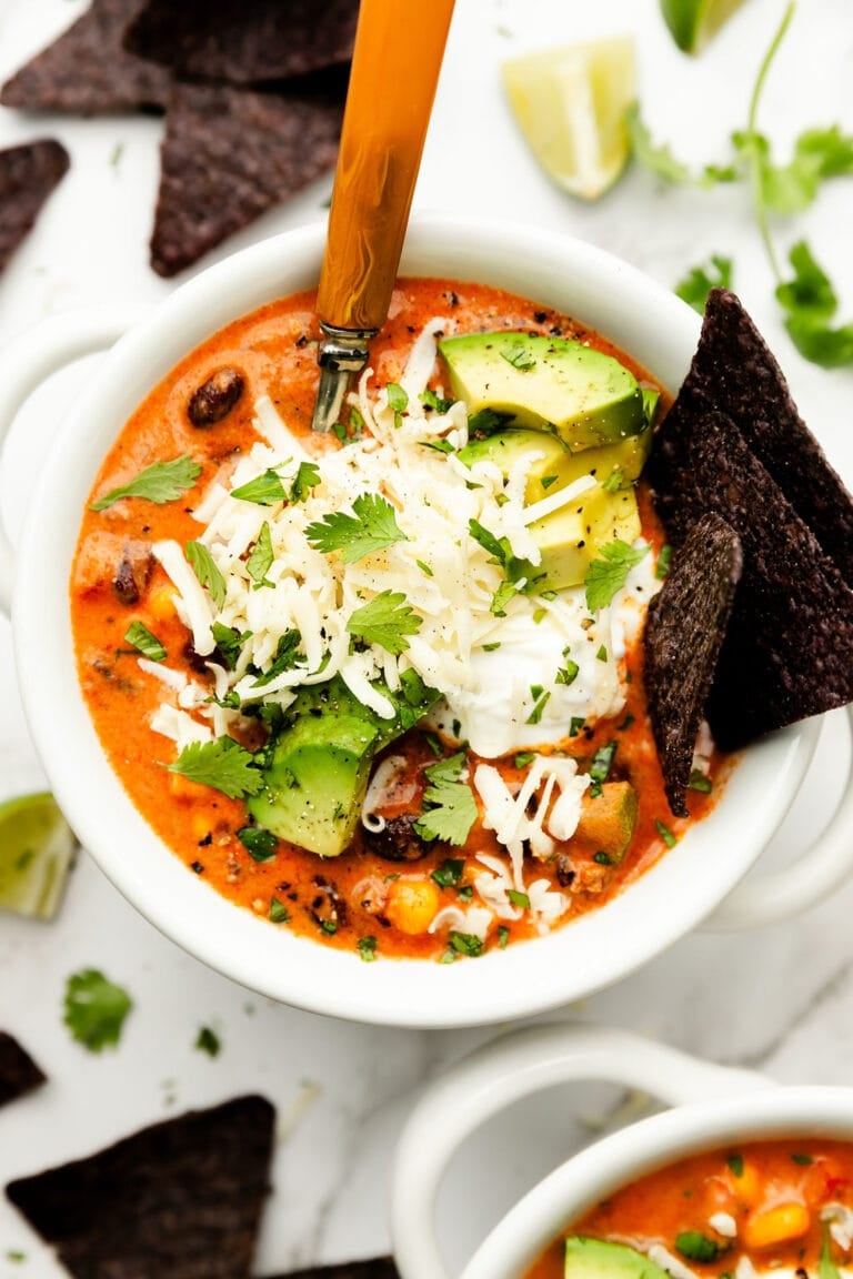Overhead view white bowl filled with taco soup, topped with sour cream, avocado pieces and blue corn chips. 
