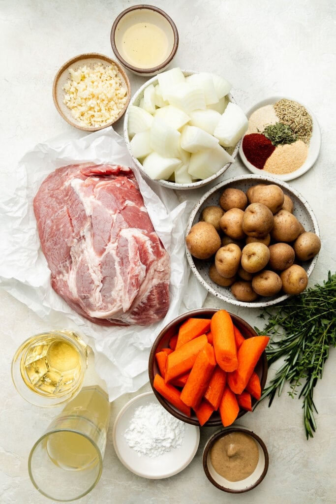 The ingredients that will be used to make crock pot pork roast, including pork shoulder, potatoes, carrots, spices, onions, minced garlic, Dijon mustard, broth, and white wine. 