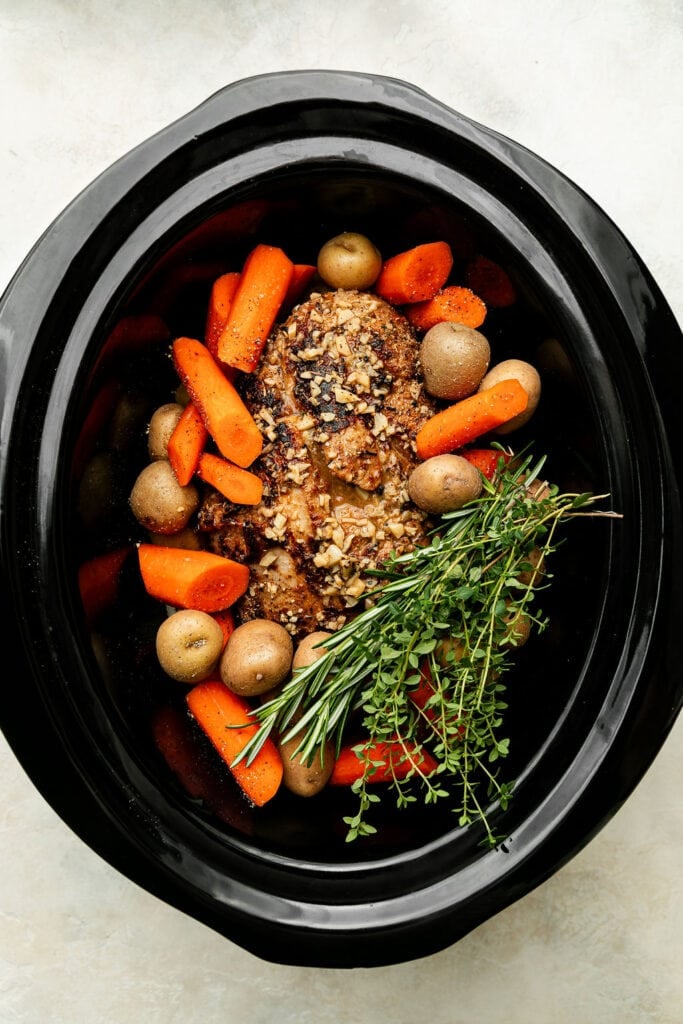 Overhead view of vegetables and a seasoned covered roast in a black crockpot garnished with a bundle of fresh herbs. 