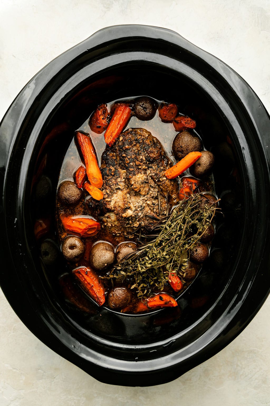 Overhead view of a seared pork butt with carrots, onions and fresh herbs in a black slow cooker insert after cooking. 