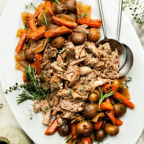 White platter filled with crockpot pork roast shredded with roasted potatoes and carrots on either ends of platter