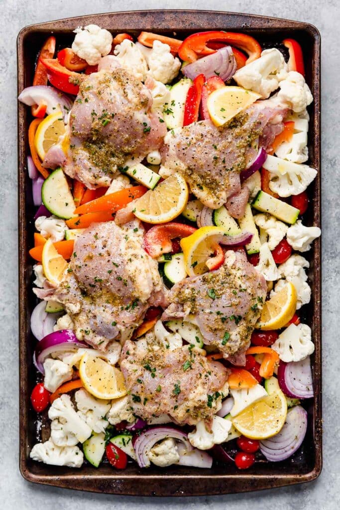 Overhead view sheet pan filled with vegetables and chicken thighs rubbed in Mediterranean spices.