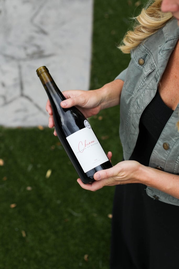 A women wearing black dress and jean jacket holding a bottle of wine with Dry Farm Wines label over stone walkway