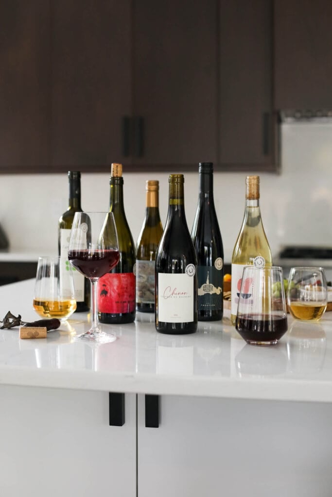 Several wine bottles from Dry Wine Farms on a white countertop, four wind glasses with small amount of wine in each around bottles.