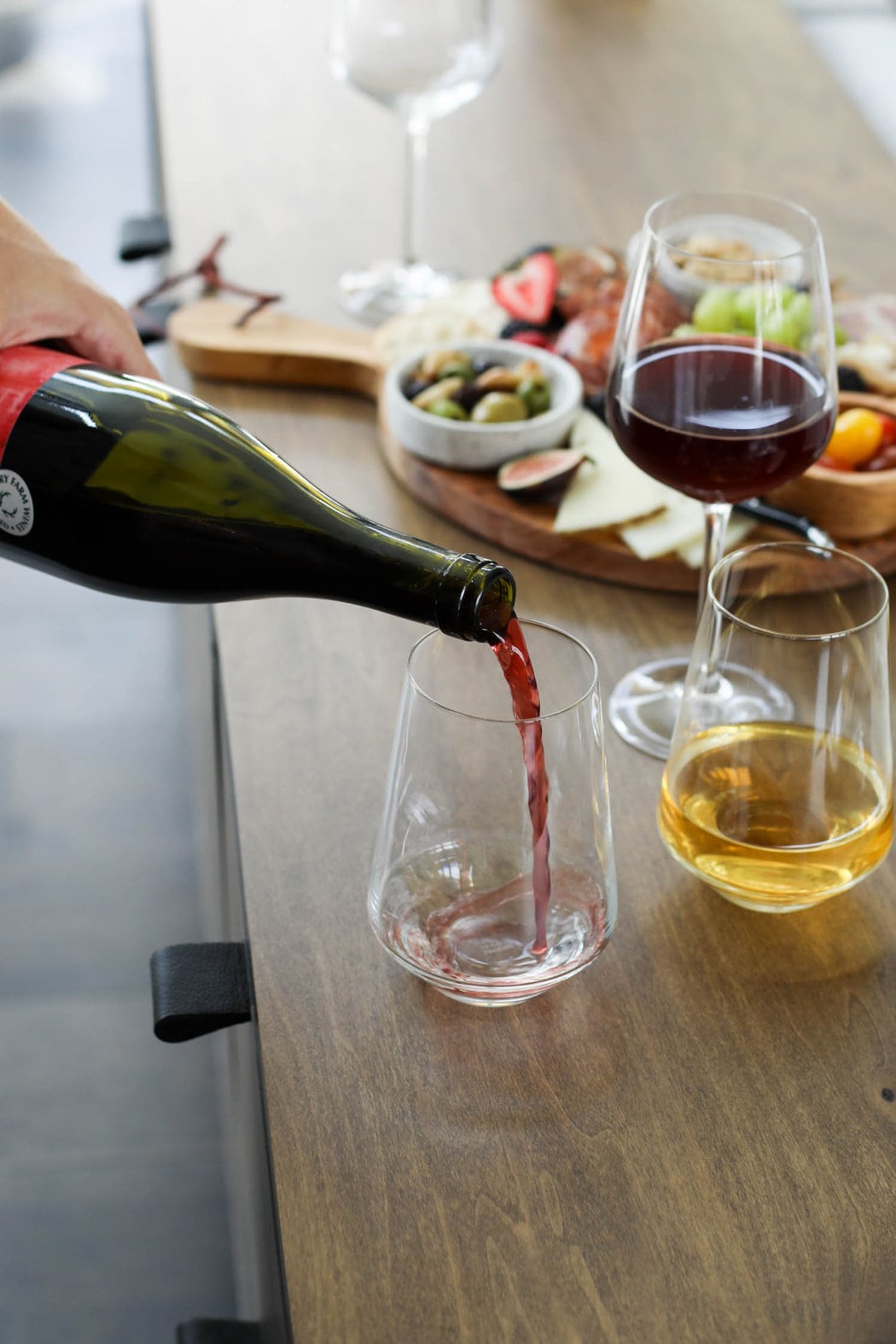 A hand pouring red wine into a stemless glass in the forefront with a charcuterie board and other glasses of wine in the background.