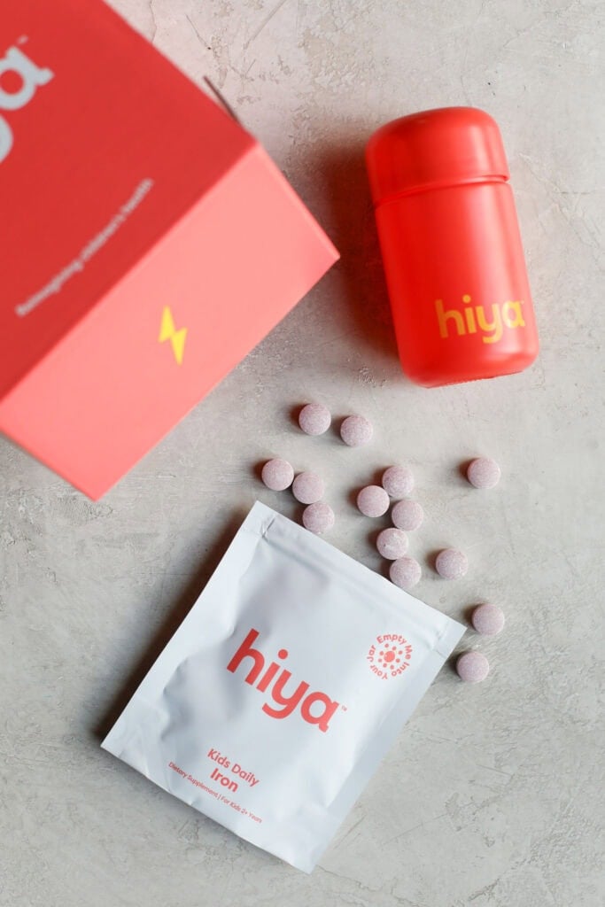 Red box packaging for Hiya kids daily iron plus supplement on countertop with individual tablets coming out of the packaging. 