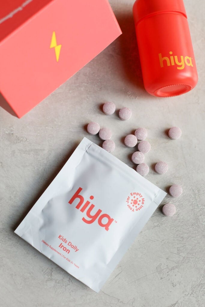 Red box packaging for Hiya kids daily iron plus supplement on countertop with individual tablets coming out of the bag. 