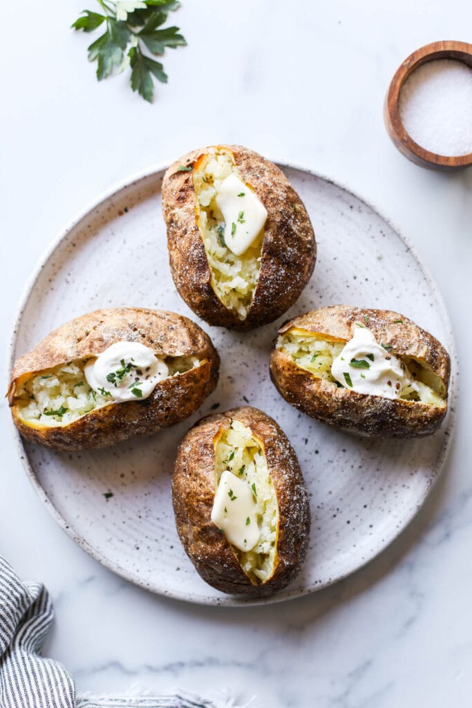 Four perfectly baked potatoes on a plate, two with sour cream and chives, and the other two with butter and parsely. 