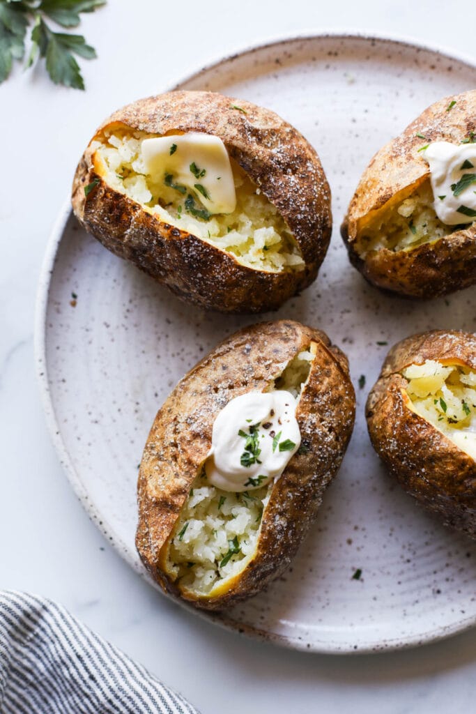 Two baked potatoes with crispy salted skin, one with butter and parsley and the other with sour cream on a plate. 