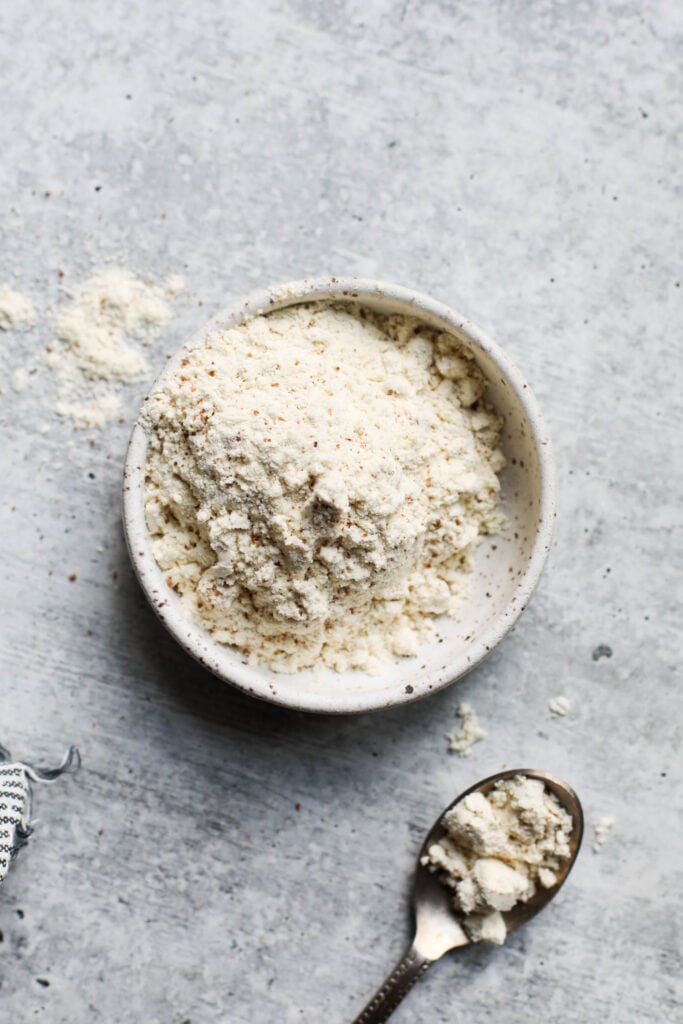 Close up view of protein powder in a small white bowl with a spoon beside it full of a scoop of the powder. 
