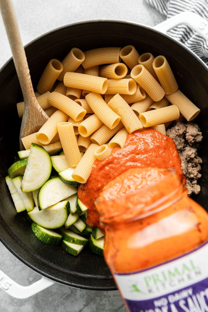 Vodka sauce being poured over rigatoni noodles, zucchini, and sausage in skillet