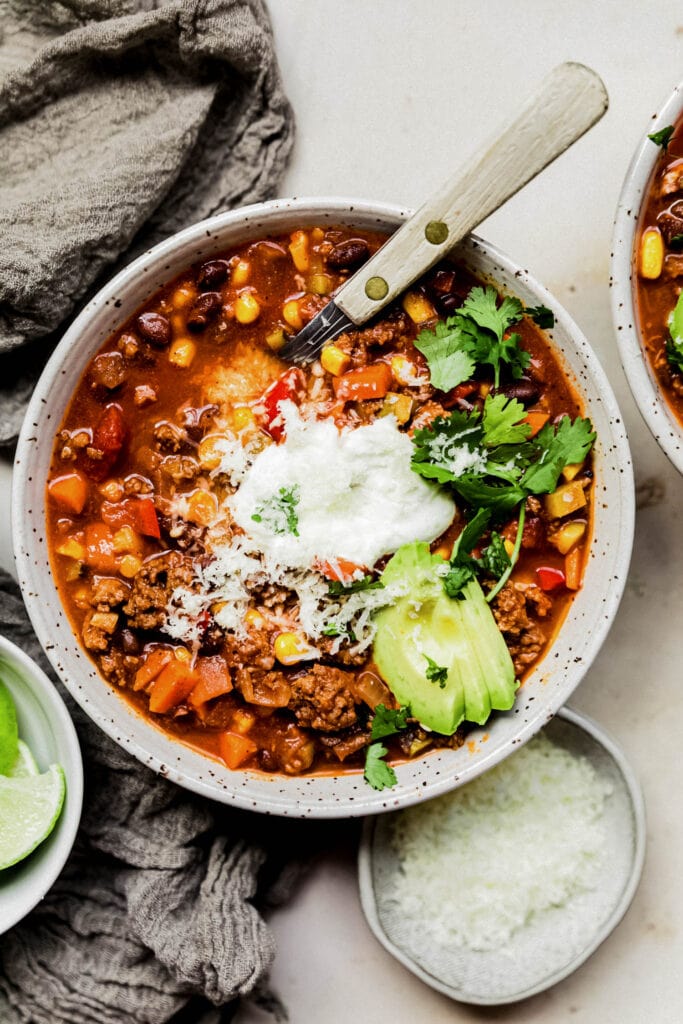 An overhead view of a bowl of healthy turkey chili topped with sour cream, avocado, cilantro, and shredded cheese. 