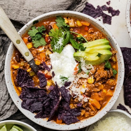 A hearty bowl of healthy turkey chili with avocado slices, sour cream, cheese, blue corn chips, and cilantro on top.