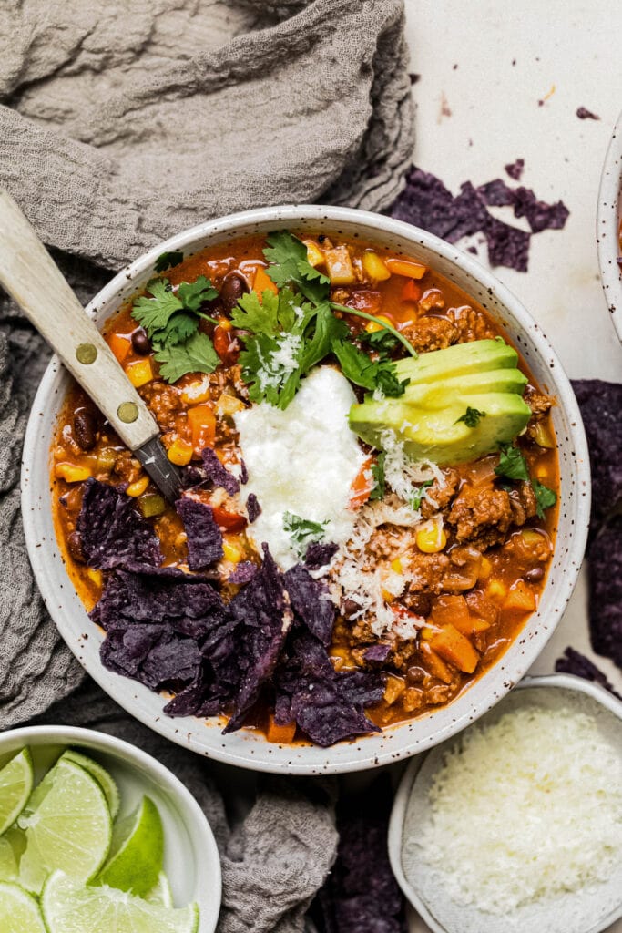 A hearty bowl of healthy turkey chili with avocado slices, sour cream, cheese, blue corn chips, and cilantro on top. 