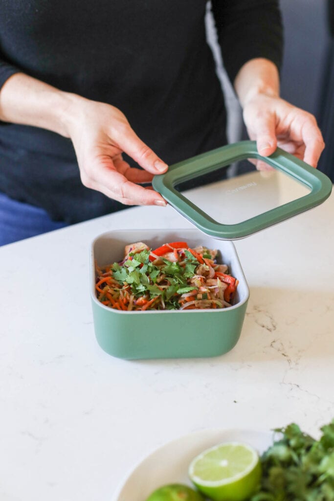 A green and glass lid being paced on a Caraway storage meal prep container filled with chicken pad thai leftovers