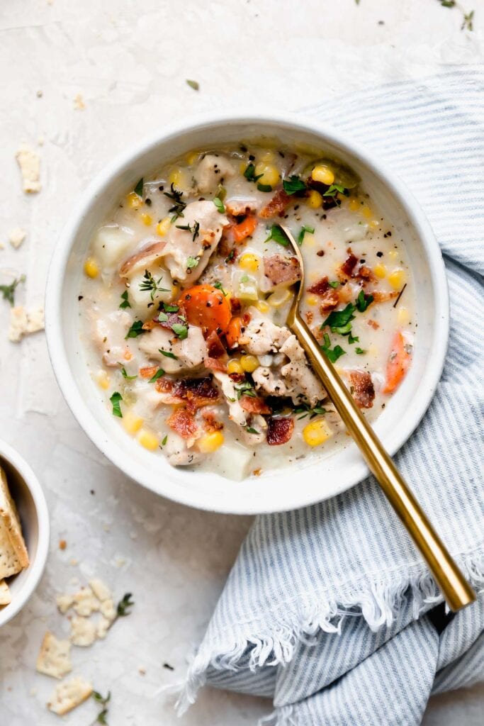 Overhead view white bowl filled with creamy chicken corn chowder, topped with crisp cooked bacon pieces, gold spoon in soup