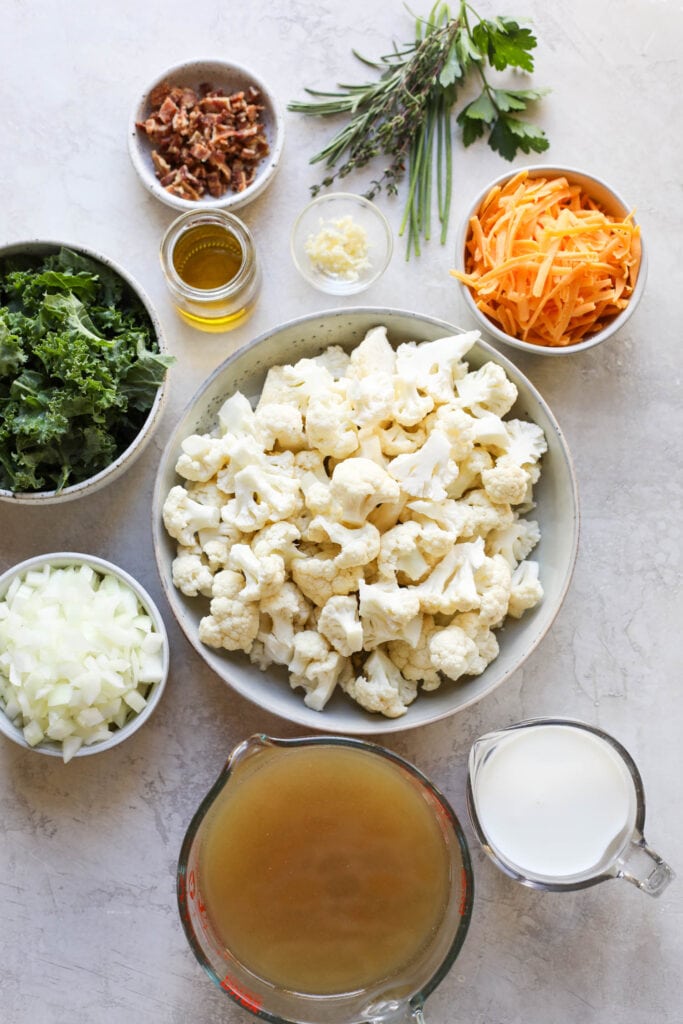 An overhead view of the ingredients to make Cauliflower Soup, including chopped cauliflower, kale, whole milk, chicken broth, onions, and herbs.