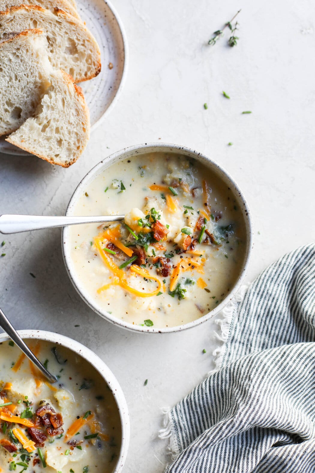 An overhead view of a bowl of Cauliflower Soup and topping on a counter with bread and another bowl of soup partially visible in the corner.