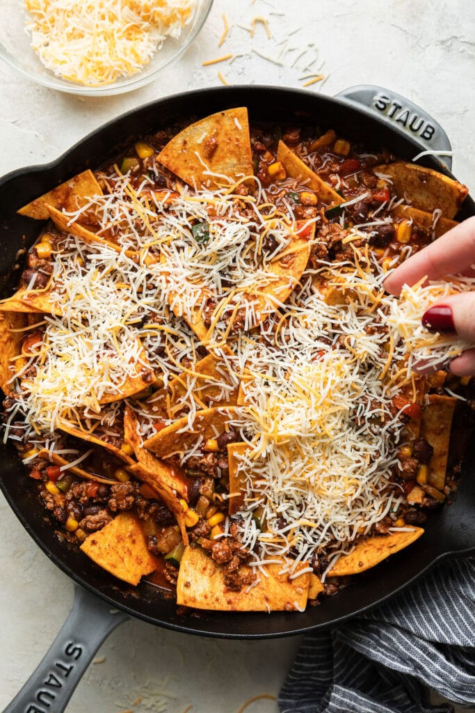 Shredded cheese being sprinkled over top of of skillet filled with beef enchiladas